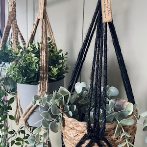 May include: Two macrame plant hangers, one made with natural jute rope and the other with black rope. Both hangers are holding potted plants. The jute hanger has a tag that reads "Handmade by Helen at Melons & Mangoes".