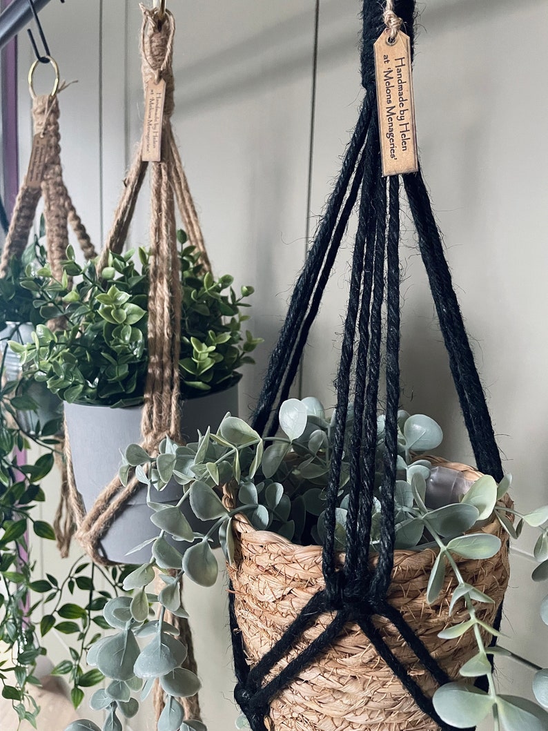 May include: Two macrame plant hangers, one made with natural jute rope and the other with black rope. Both hangers are holding potted plants. The jute hanger has a tag that reads "Handmade by Helen at Melons & Mangoes".