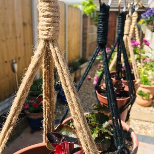 May include: Three hanging plant pots with macrame plant hangers. The pot in the foreground is brown with a dark red plant. The other two pots are black with green plants. The hangers are made of natural jute rope and black cord.