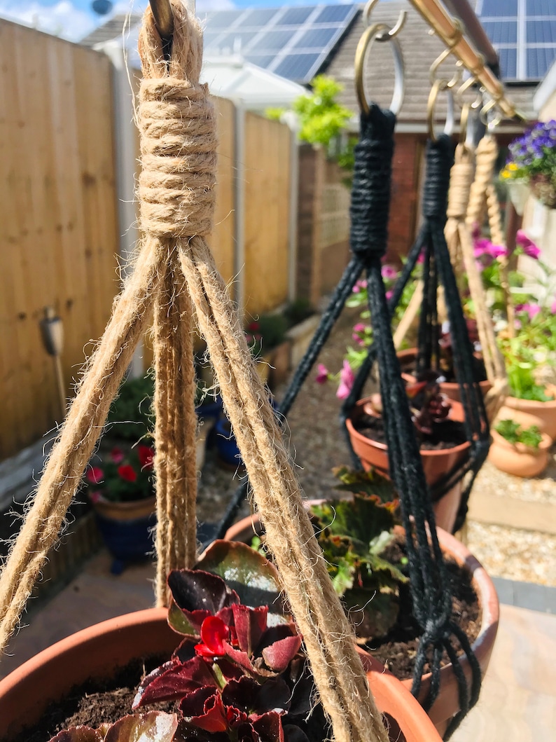 May include: Three hanging plant pots with macrame plant hangers. The pot in the foreground is brown with a dark red plant. The other two pots are black with green plants. The hangers are made of natural jute rope and black cord.