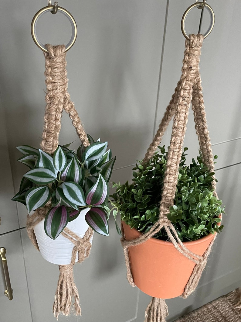 May include: Two macrame plant hangers with pots and plants. The plant hangers are made of natural jute rope and have a simple design. One plant is in a white pot and the other is in a terracotta pot.