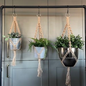 May include: Three macrame plant hangers with different colored pots hanging from a black metal rod. The hangers are made of natural jute rope and have a tassel at the bottom. The pots are filled with green plants.