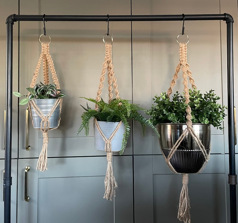 May include: Three macrame plant hangers with different colored pots hanging from a black metal rod. The hangers are made of natural jute rope and have a tassel at the bottom. The pots are filled with green plants.