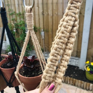 May include: A close-up of a natural fiber macrame plant hanger. The hanger is made of two strands of thick, natural-colored rope. The rope is knotted in a complex pattern to create a sturdy and decorative hanger. The hanger is attached to a metal ring at the top.