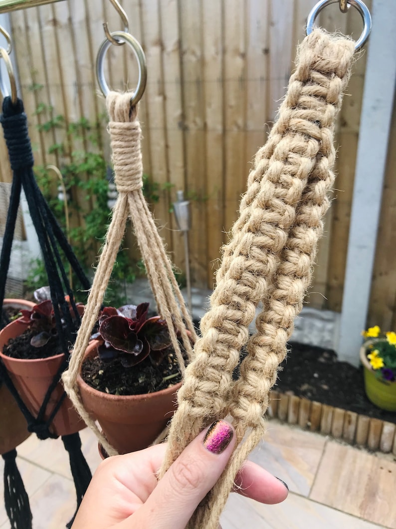 May include: A close-up of a natural fiber macrame plant hanger. The hanger is made of two strands of thick, natural-colored rope. The rope is knotted in a complex pattern to create a sturdy and decorative hanger. The hanger is attached to a metal ring at the top.