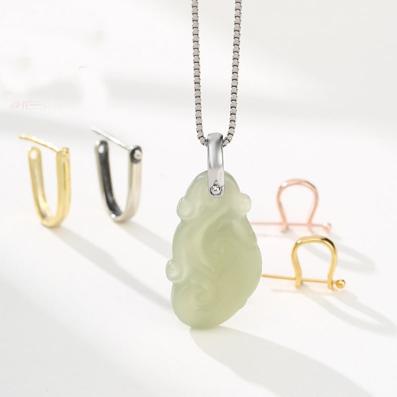 May include: A silver chain necklace with a jade pendant in the shape of a mythical creature. The pendant is carved with intricate details and has a small diamond at the top. The necklace is displayed with three different types of clasps.