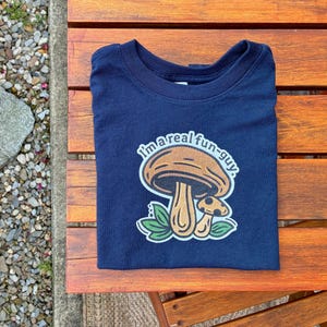 May include: Navy blue t-shirt with a mushroom graphic and the text "I'm a real fun-guy." The shirt is folded on a wooden surface next to a garden bed with small stones and green foliage.