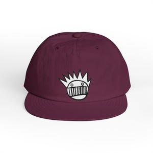 May include: A deep maroon baseball cap featuring a centered white embroidered graphic of a stylized circular face with spiky hair and a wide, toothy grin. The cap has a structured crown, a flat brim, and a button detail at the top center.