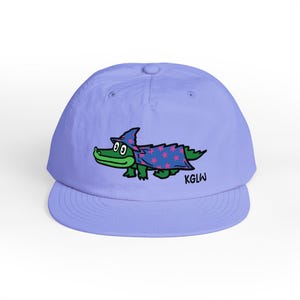 May include: A light purple baseball cap featuring a cartoon alligator wearing a wizard's robe with pink stars. The alligator has green skin and is wearing a blue hat. The text "KGLW" is printed on the right side of the cap.