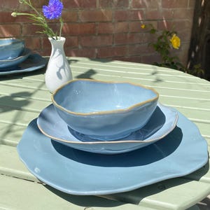 May include: A set of light blue ceramic tableware with a wavy edge, comprising a large plate, a smaller plate, and a bowl. The set is on a green table. A small white vase holds a blue flower.