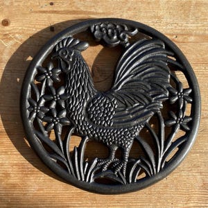 May include: Black cast iron trivet featuring a rooster design. The rooster is surrounded by flowers and leaves. The trivet is round and has a diameter of approximately 20 cm.