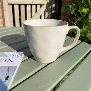 May include: A pale cream ceramic mug with a curved handle rests on a green slatted table. The mug has a light brown rim and is next to a magazine with the text "2025 DECORATING GUIDE". A wooden fence and foliage are in the background.