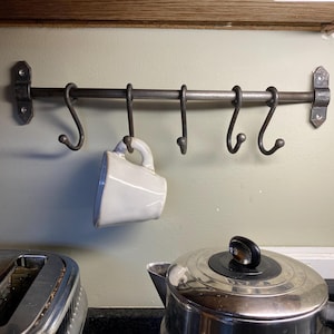 Peut inclure: Un support de cuisine en fer forgé avec six crochets, monté sur un mur vert clair. Une tasse blanche est suspendue à l'un des crochets. Une bouilloire et un grille-pain en acier inoxydable sont au premier plan.