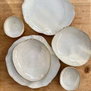 May include: Set of white ceramic bowls and plates with a slightly rippled edge and a thin gold rim. The bowls and plates are arranged on a wooden surface.