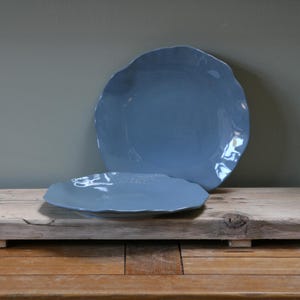 Könnte beinhalten: Zwei blau-graue Keramikplatten mit organischen, wellenförmigen Rändern. Die Platten werden auf einer rustikalen Holzoberfläche präsentiert, wobei eine größere Platte hinter einer kleineren liegt. Die Platten haben eine matte Oberfläche und ein schlichtes, elegantes Design.