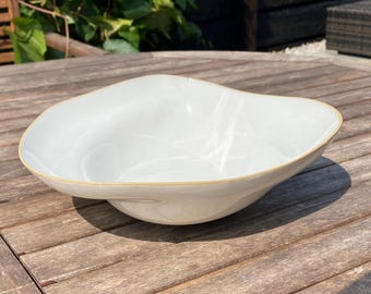 May include: A white, organically shaped bowl with a gold rim. The bowl has a smooth, glossy finish and sits on a wooden surface. The bowl's design is modern and elegant, suitable for serving or display.