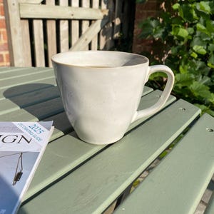 May include: A pale cream ceramic mug with a curved handle, filled with a light-coloured liquid, sits on a light green wooden table. A magazine with the text "2025 DECORATING GUIDE" is next to the mug. The background features a wooden fence and foliage.