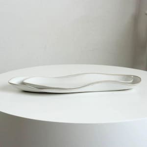 May include: Two white ceramic serving platters with a wavy design. The platters are nested together, with a smaller one inside a larger one. The edges have a subtle, light-coloured trim. The platters are on a white surface.