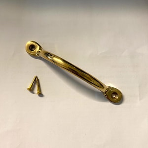 Könnte beinhalten: Ein polierter Messing-Schrankgriff mit zwei passenden Schrauben. Der Griff hat eine gebogene Stangenform mit Befestigungslöchern an jedem Ende. Die Hardware hat eine goldglänzende Farbe.