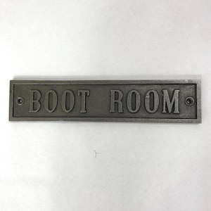 May include: A rectangular, gray metal sign with the words "BOOT ROOM" embossed in capital letters. The sign has a raised border and two small holes, one on each end. The sign is designed to be mounted on a wall.
