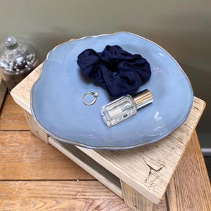 May include: A pale blue ceramic dish with a gold edge contains a navy scrunchie, a gold ring, and a small perfume bottle. The dish rests on a wooden surface. A glass jar with a lid is in the background.
