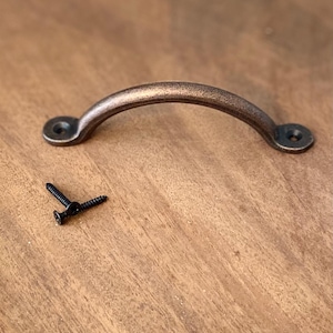 May include: A rustic, antique-style,  bronze-colored metal cabinet pull with a rounded, arched shape. Two black screws are included.