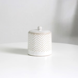May include: A white ceramic jar with a lid. The jar is cylindrical and decorated with a chevron pattern of small, raised dots. The lid has a small, round knob. The jar is on a white surface. The jar is approximately 10 cm in height.