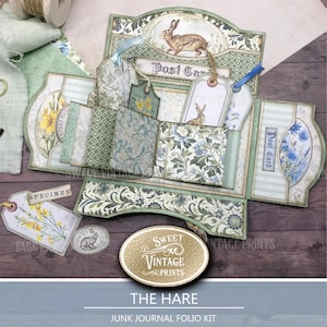 Può includere: Un kit di folio di diario spazzatura intitolato "The Hare" con un'estetica vintage. Il kit presenta una scatola decorativa con illustrazioni floreali e di conigli, etichette e tasche. La tavolozza dei colori include sfumature di verde, blu e crema, con il testo "Post Card" e "Specimen" visibili.