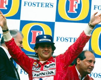 May include: A race car driver wearing a red racing suit with the words "Marlboro McLaren International" and "Honda" on the front, raises both arms in victory. The driver is wearing a blue baseball cap with the word "Nacional" on it. The driver is standing in front of a wall with the word "Foster's" repeated on it.