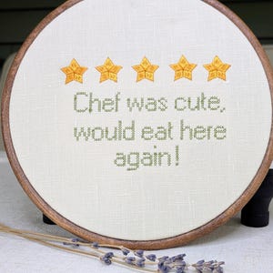 May include: A circular wooden embroidery hoop containing off-white fabric with five embroidered yellow stars and the green stitched text: Chef was cute, would eat here again! The design is centred on the fabric within the dark brown wooden frame.