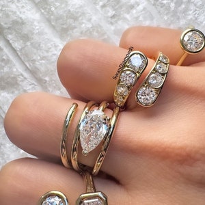 Könnte beinhalten: Eine Sammlung von Goldringen mit verschiedenen Diamantfassungen. Die Ringe haben einen birnenförmigen Diamanten, einen runden Diamanten und einen quadratischen Diamanten. Die Ringe sind auf einer Hand gestapelt.