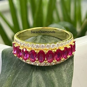 Può includere: Anello a fascia d'oro con una fila di pietre ovali rosa e una fila di pietre trasparenti più piccole. L'anello è su una foglia verde.