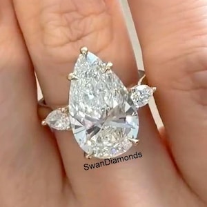 Peut inclure: Une bague en or ornée d'un diamant en forme de poire, flanqué de deux diamants plus petits. Le nom de la marque "SwanDiamonds" est visible sur la bague.