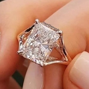 Puede incluir: Un anillo de compromiso de oro rosa con un gran diamante rectangular transparente. El diamante está engastado en una banda delicada y calada con dos garras a cada lado. El anillo está en un dedo con esmalte de uñas rosa claro.