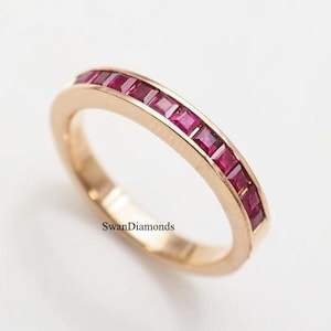May include: A gold ring featuring a line of square-cut, deep red gemstones. The ring has a polished finish and the text "SwanDiamonds" is visible on the band.