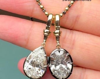 May include: A gold necklace featuring two diamond pendants. One pendant is pear-shaped, the other is oval. The diamonds are set in black metal and hang from gold clasps. The chain is delicate, with small gold beads. The text "SwanDiamonds" is visible.
