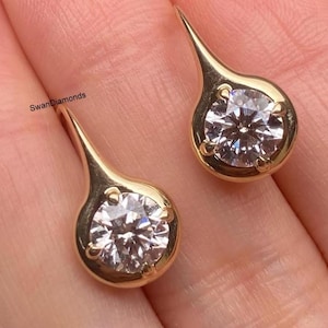 May include: A pair of gold-colored teardrop earrings, each featuring a round, sparkling diamond. The earrings are set in a bezel setting, with a slender, elongated hook. The brand name "SwanDiamonds" is visible on the left earring.