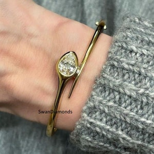 Puede incluir: Una pulsera dorada con un diamante en forma de pera. La pulsera tiene un diseño abierto único que se envuelve alrededor de la muñeca. El diamante está engastado en un bisel. La pulsera se muestra en una muñeca, con un suéter gris en el fondo.