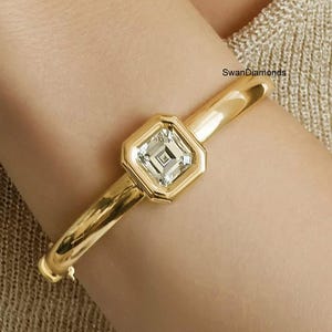 May include: A gold-colored bangle bracelet with a square, clear gemstone at the center. The gemstone is set in a gold bezel with an octagonal shape. The bracelet is smooth and shiny, with the text "SwanDiamonds" visible.