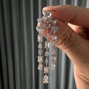 Puede incluir: Elegantes pendientes de diamantes con un diseño en cascada. Los pendientes presentan una variedad de formas de diamantes, incluyendo corazones, cortes esmeralda y formas de pera. Los pendientes están engarzados en un metal plateado. La marca "SwanDiamonds" es visible.