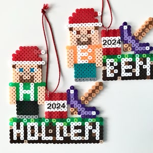 May include: Pixelated Christmas ornaments featuring figures wearing Santa hats. One ornament says "HOLDEN" and the other says "BEN", both with the year "2024". The ornaments are made of colourful beads and have red hanging strings.
