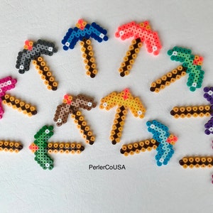 May include: A collection of colorful pixel art pickaxes in various colors, including gray, blue, pink, green, brown, yellow, and purple. Each pickaxe features a brown handle and a contrasting head, made from small, fused plastic beads. The text "PerlerCoUSA" is visible.