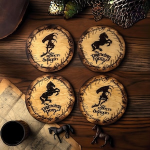 May include: Four circular wooden coasters with a light brown finish. Each coaster features a black silhouette design of a dragon or a horse, with the words "The Green Dragon" or "The Prancing Pony" in a decorative font. The coasters are arranged on a wooden surface.