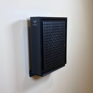 Puede incluir: Un purificador de aire negro montado en una pared blanca. El dispositivo tiene forma rectangular con un panel frontal enrejado. La marca "hitron" es visible en el lateral.