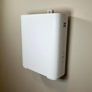 May include: A white, rectangular device mounted on a wall. The device has rounded edges and a smooth surface. It features a small logo and several indicator lights. The device is attached to the wall with a small, white bracket.