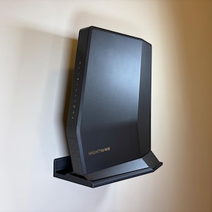 May include: A black Nighthawk router mounted on a wall. The router has a sleek, modern design with the brand name in gold lettering. It is mounted on a black shelf.
