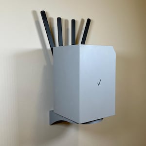 May include: A light gray router with black antennas mounted on a wall. The router has a Verizon logo and is supported by a gray shelf. The antennas are arranged vertically, extending upwards from the router.