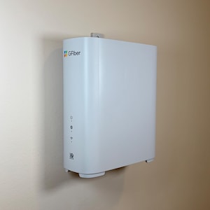May include: A white GFiber network router mounted on a wall. The device features a vertical rectangular shape with rounded edges, a multi-colored logo at the top, and four status indicator icons printed vertically along the left side panel.