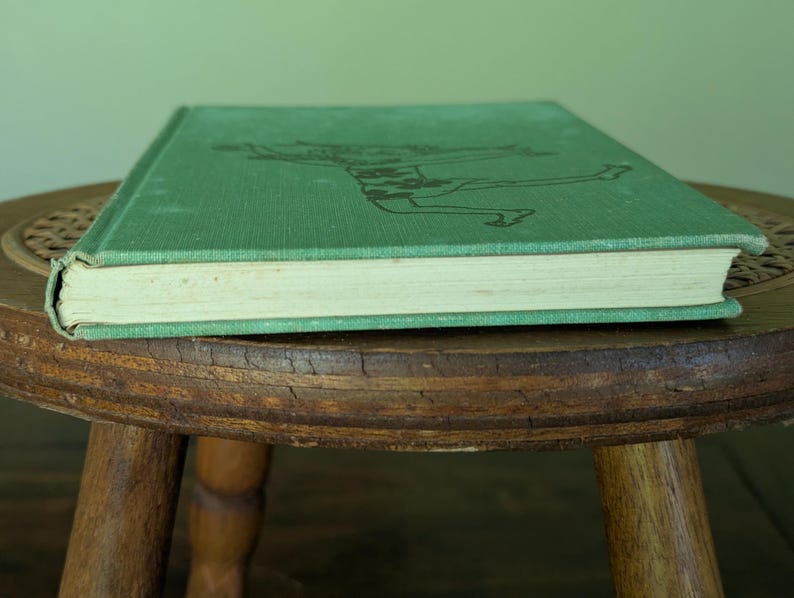 Pu&ograve; includere: Un libro vintage con copertina rigida verde e un'illustrazione sbiadita. Il libro &egrave; appoggiato su uno sgabello rotondo di legno. Le pagine sono ingiallite e il dorso del libro &egrave; visibile.