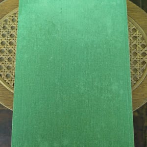 Pu&ograve; includere: Un libro con copertina rigida verde con una texture visibile. La copertina mostra segni di usura. &Egrave; appoggiato su una superficie di legno con un motivo intrecciato.